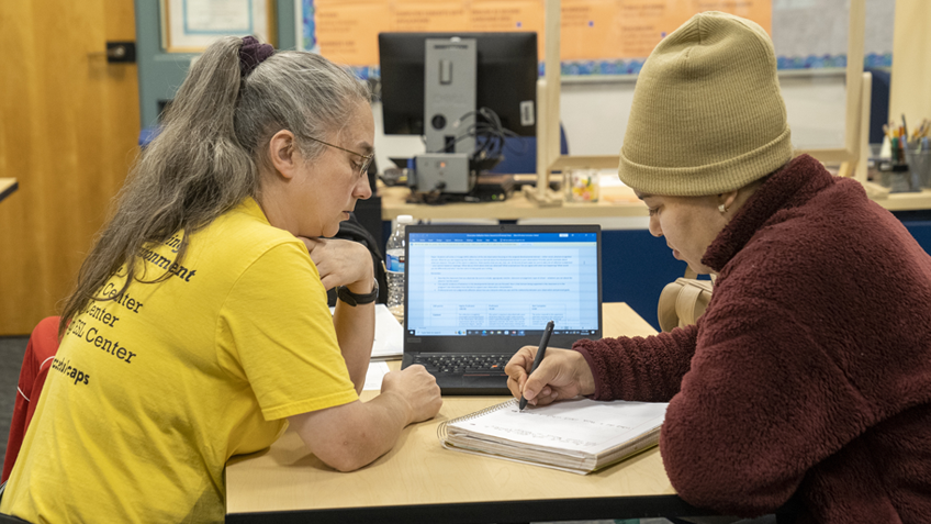 A student and a counselor sit at a table, talking over a laptop and notebook