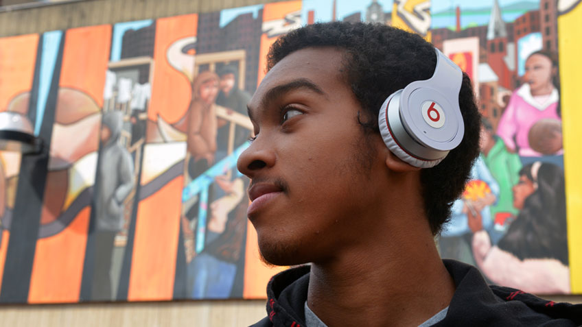 A male african american student wearing headphones stands in front of a mural